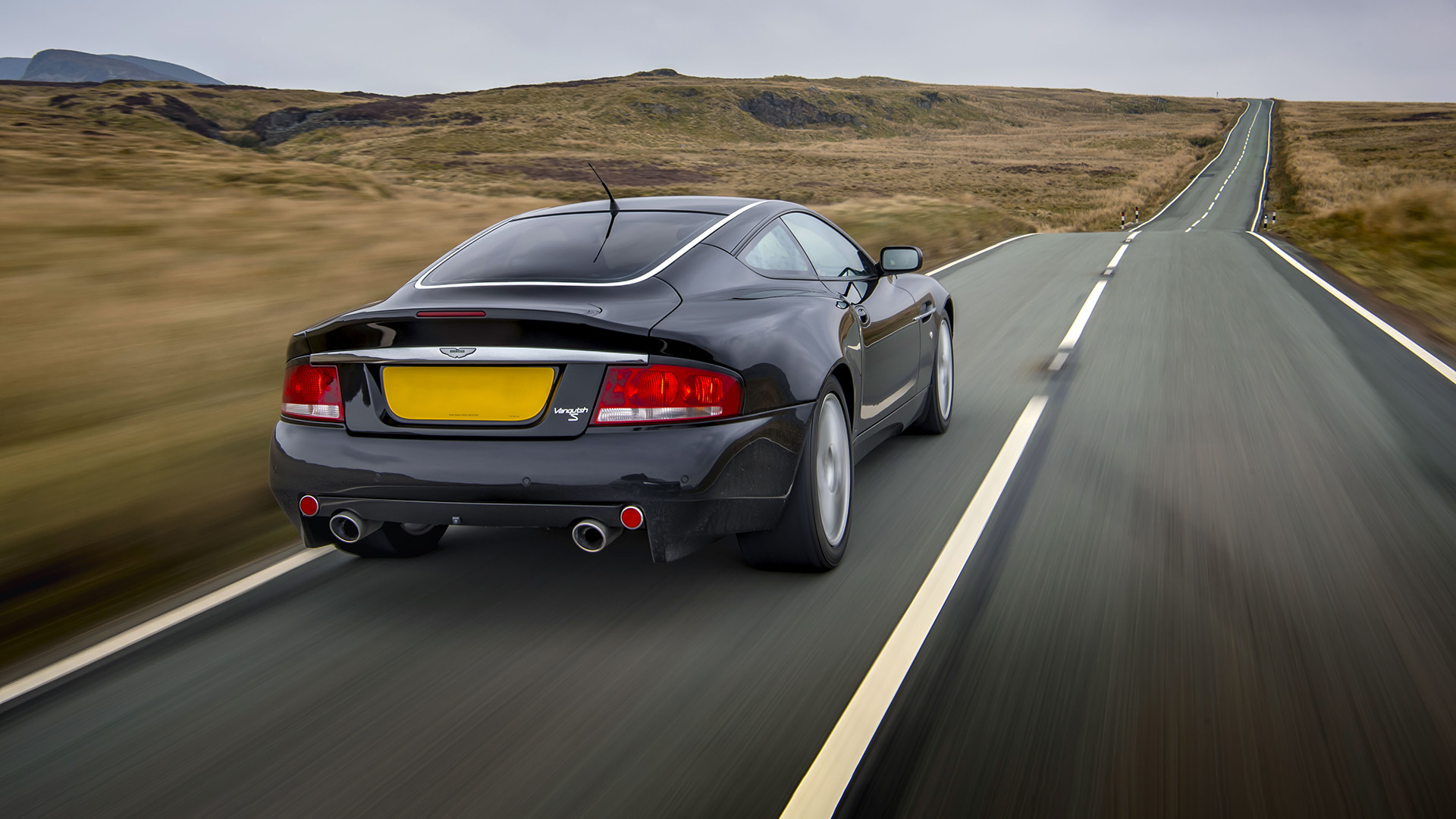 Vista trasera de un elegante Aston Martin Vanquish S negro circulando por una pintoresca carretera rural. 