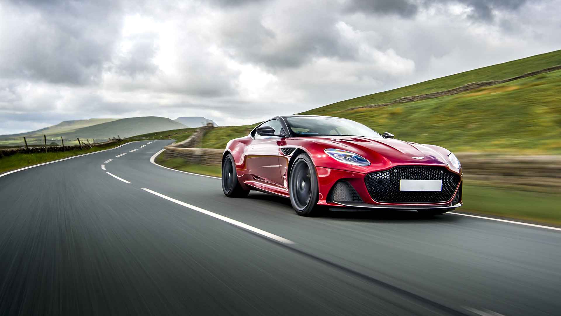 Rojo Aston Martin DBS conduciendo rápido en una carretera rural con un cielo gris arriba.