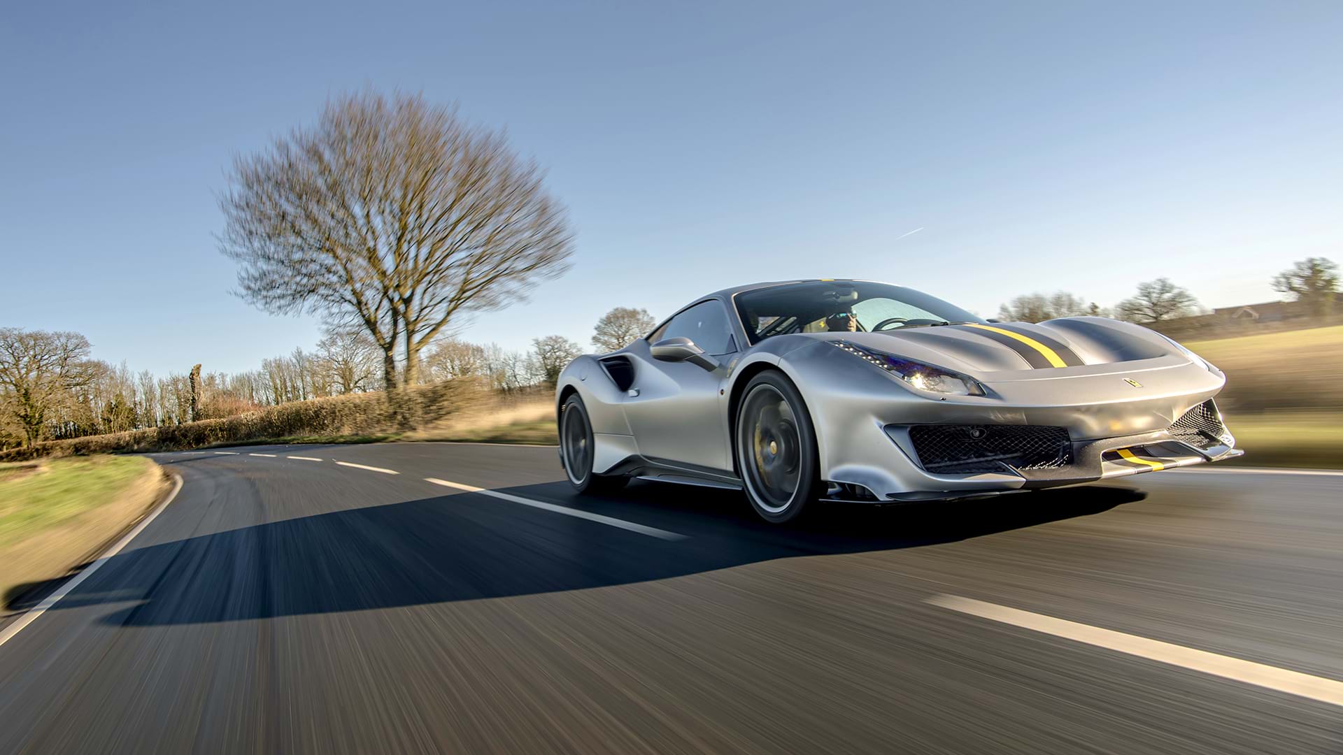 Elegante Ferrari 488 Pista gris con una llamativa franja de carreras que recorre un pintoresco paisaje campestre. 
