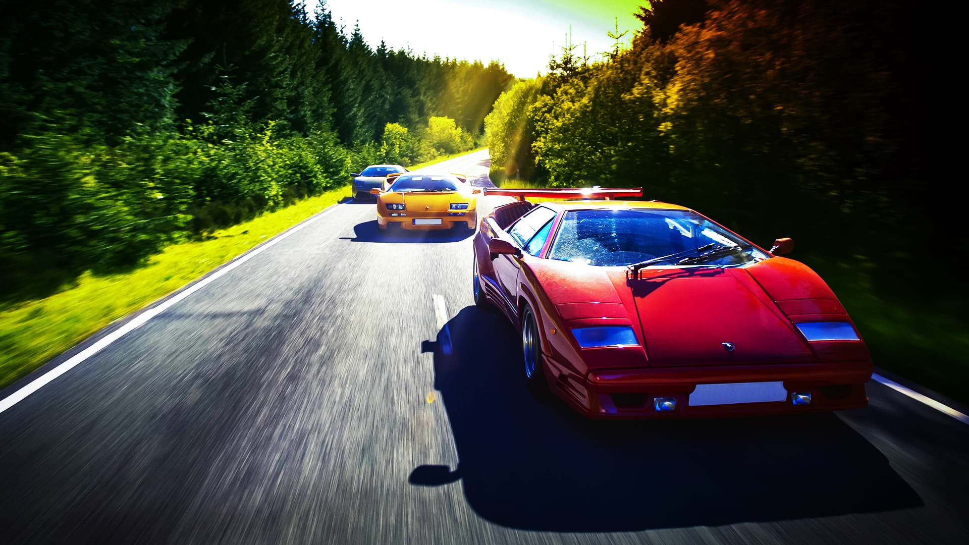 Un colorido convoy Lamborghini: Countach 25th Anniversary, Diablo 6.0 y Murciélago, corre por una carretera panorámica en un día luminoso.