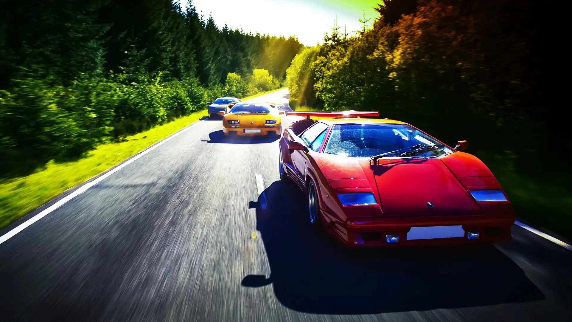 Un colorido convoy Lamborghini: Countach 25th Anniversary, Diablo 6.0 y Murciélago, corre por una carretera panorámica en un día luminoso.
