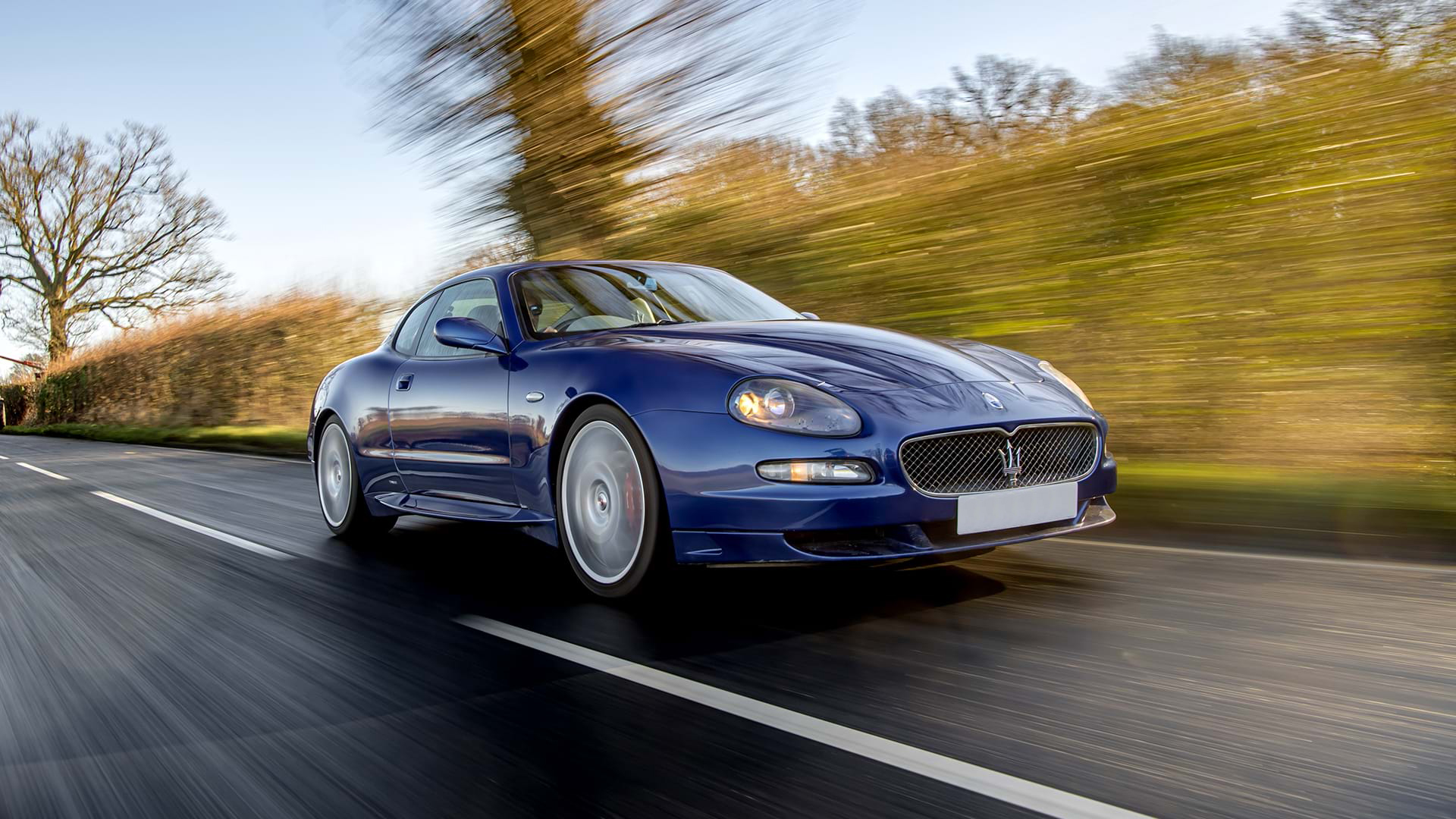 Classic Blue Maserati GranSport conduciendo por una carretera rural con cielos azules.