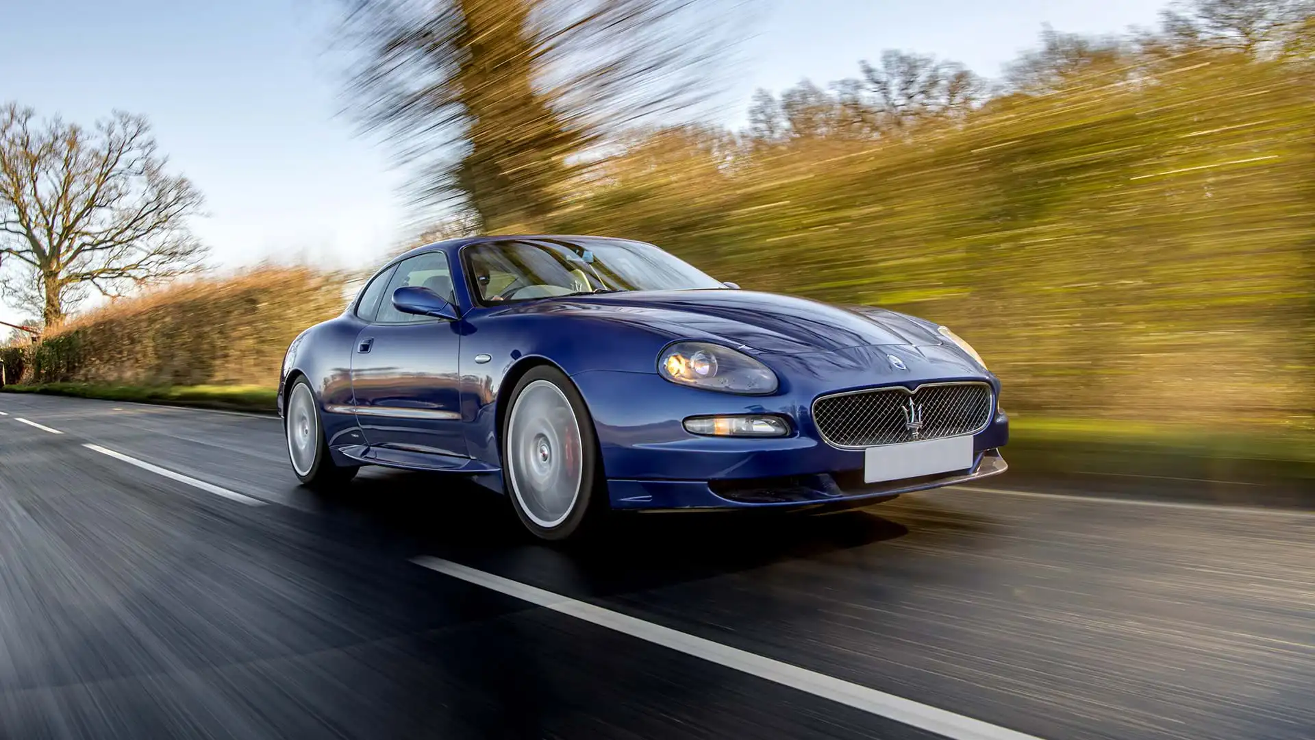 Classic Blue Maserati GranSport conduciendo por una carretera rural con cielos azules.