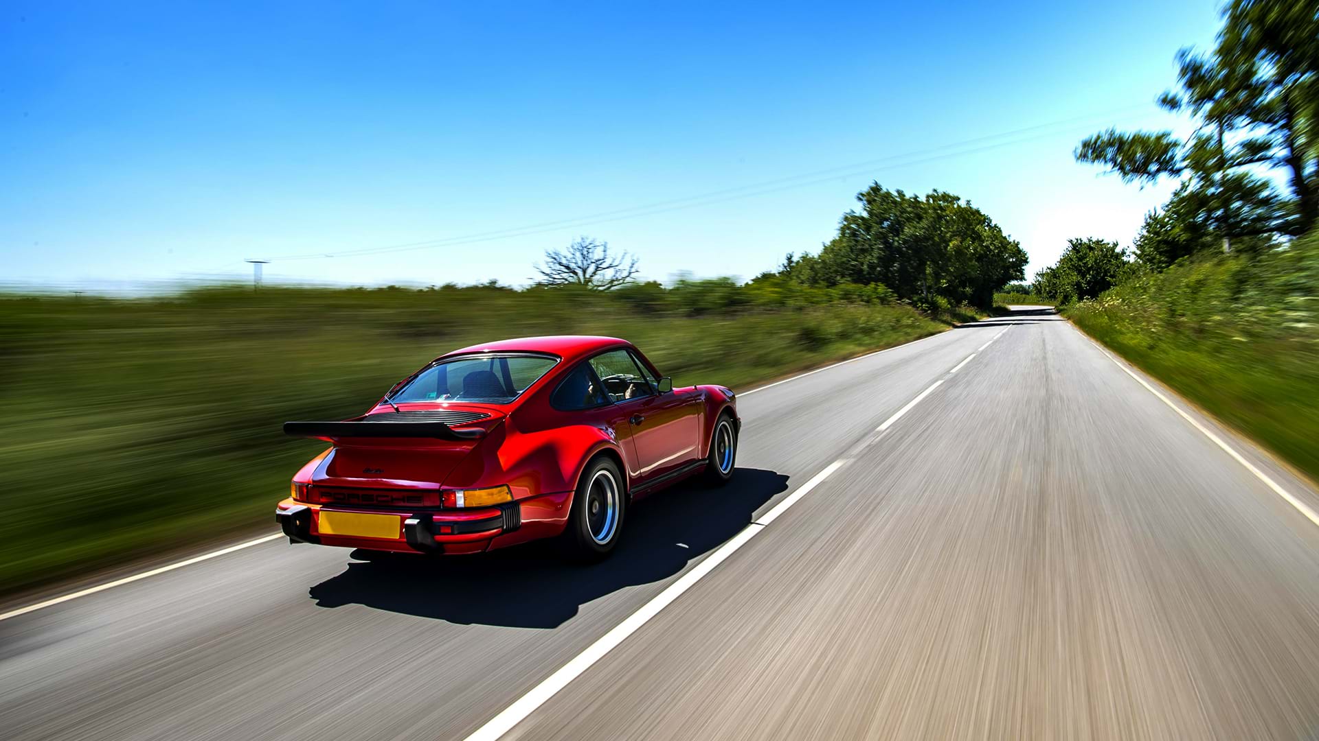 Turbo rojo vintage Porsche 930 rugiendo por un hermoso camino rural en un día soleado.
